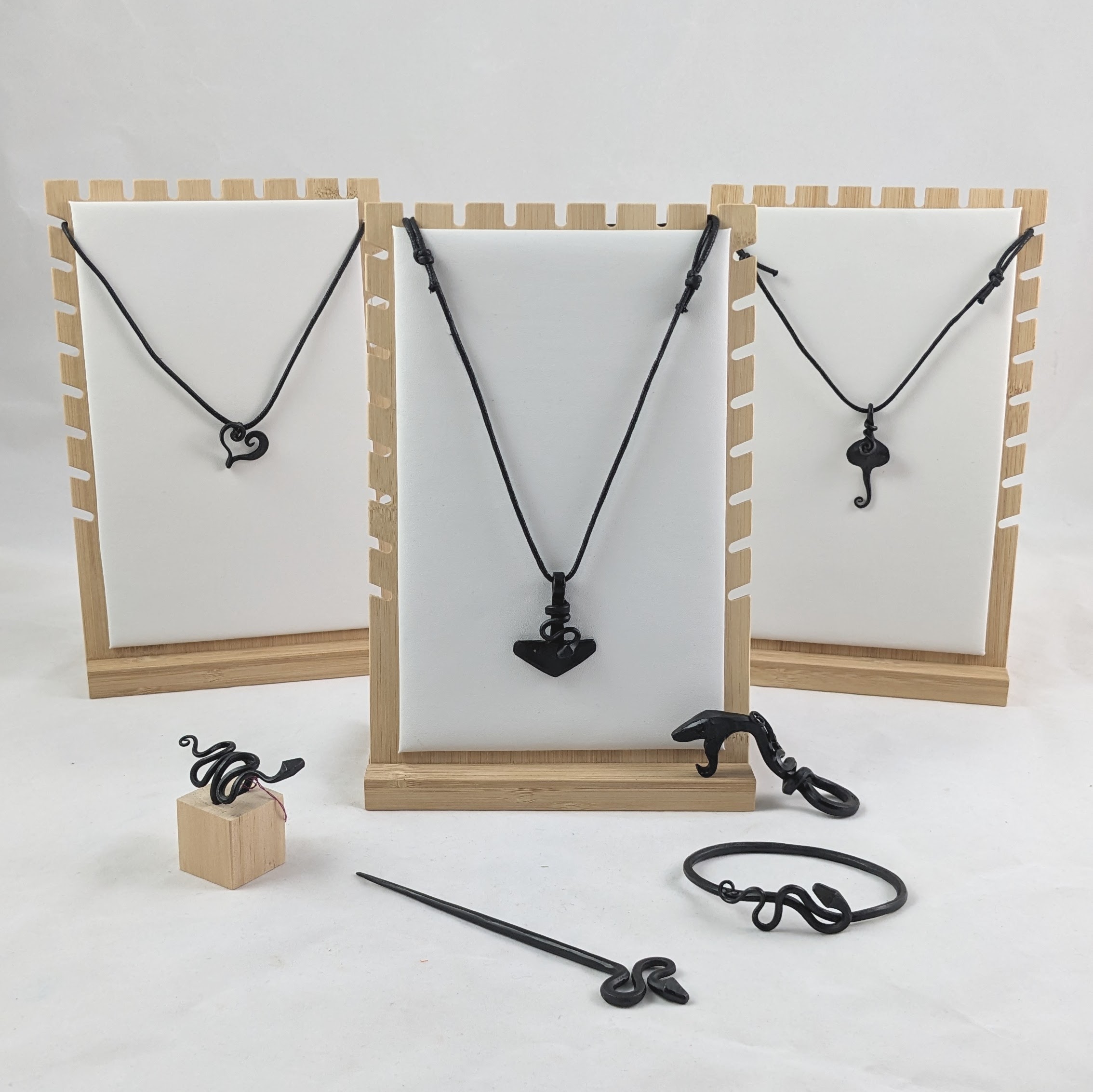Three necklace displays showing the three variations of necklaces on black leather cords: heart, thor's hammer and a heart. In the foreground is a forged snake ring, snake hairpin and cuff. 