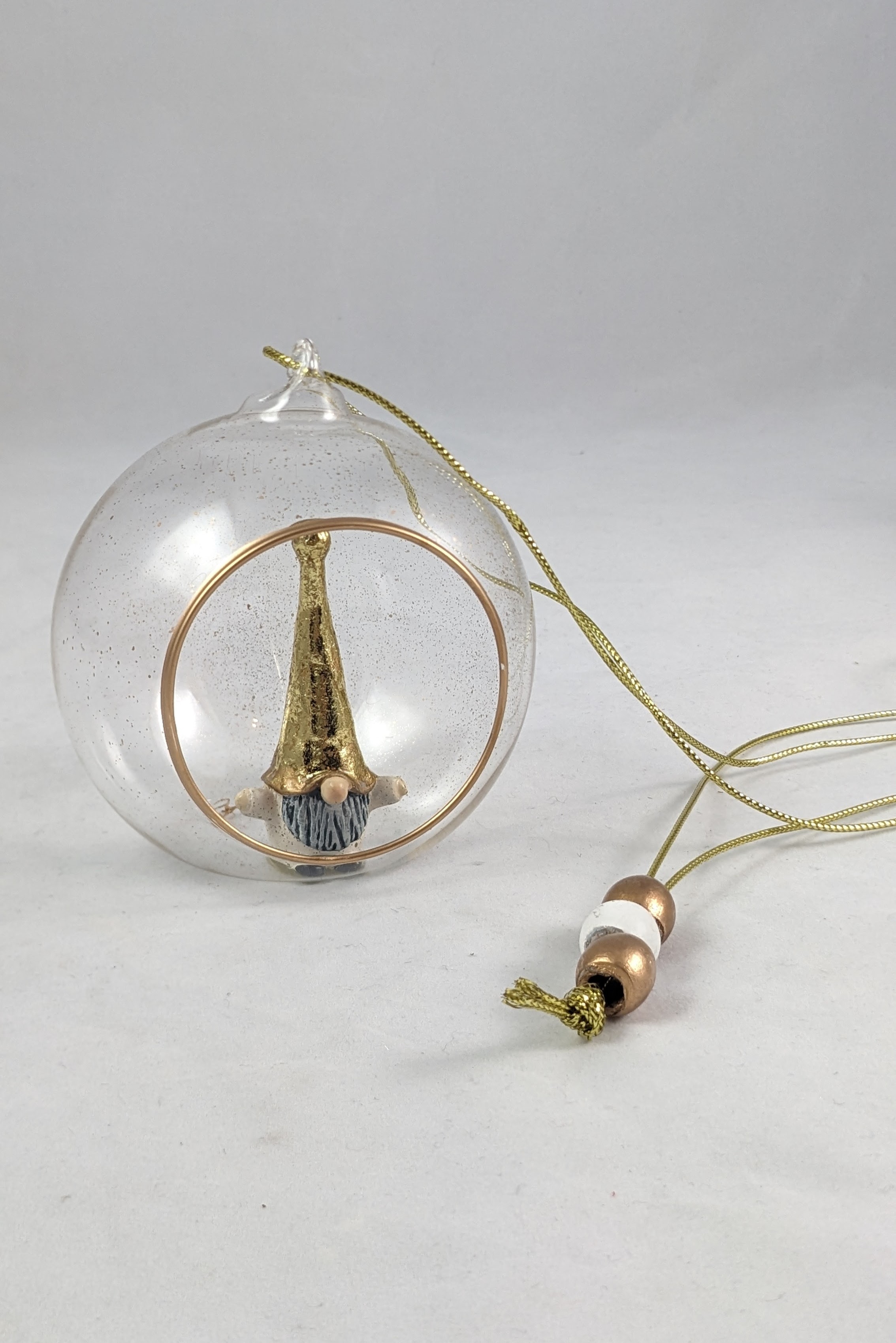 Transparent globe that has specks of gold glitter has a small opening in the front and shows a gold and white tomten inside the globe. a metallic string is connected to the the top and shows three beads on the strand. 